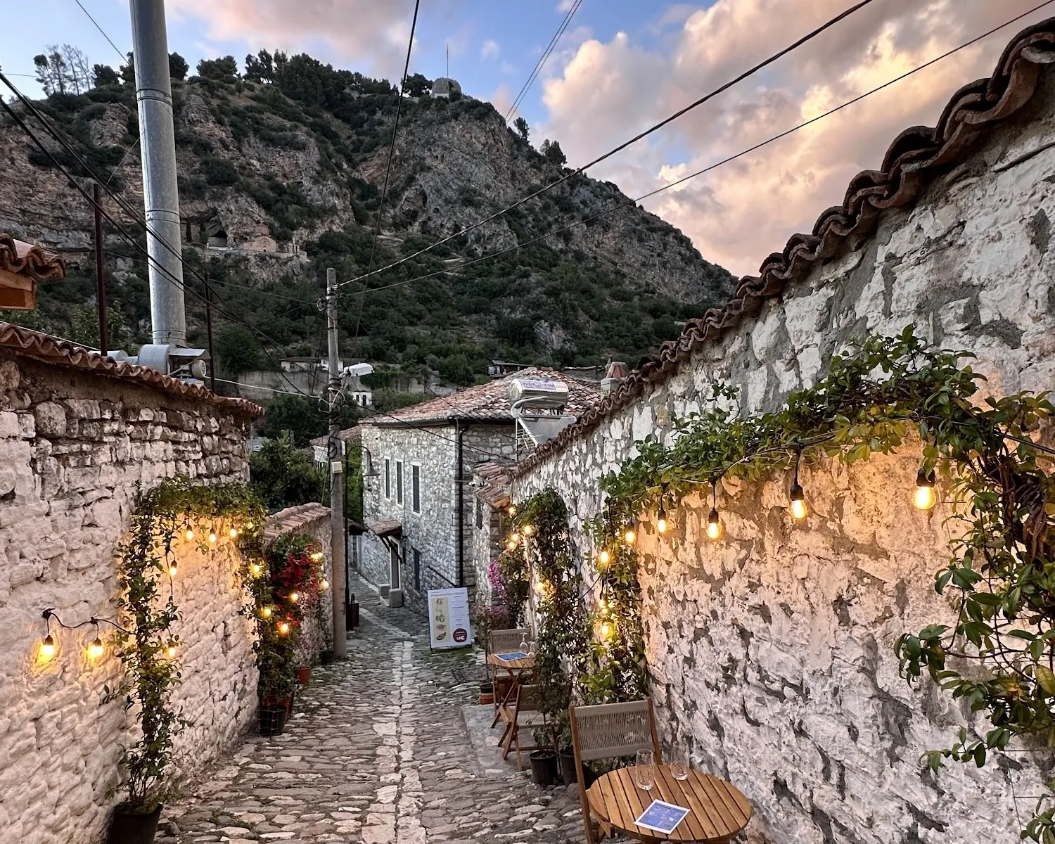 A cobbled lane in Berat's old quarter at dusk, string lights on white stone walls, small wooden tables set for two and the castle hill behind