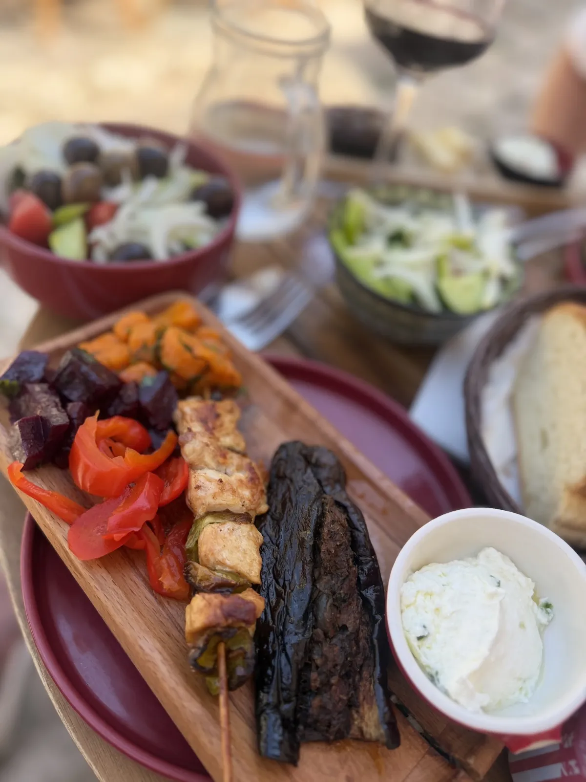 A wooden board with chicken shishkebab, stuffed eggplant, roasted peppers, pickled beetroot and carrots, and a bowl of yogurt sauce