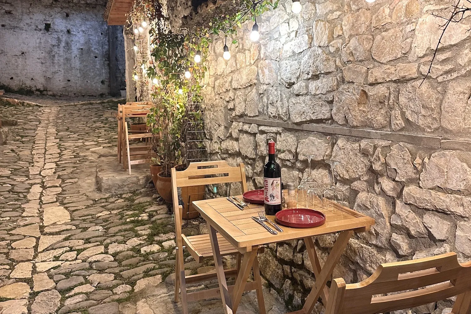 A wooden table for two against a lit stone wall at night, red plates and a bottle of Amalia red wine, on a cobbled lane