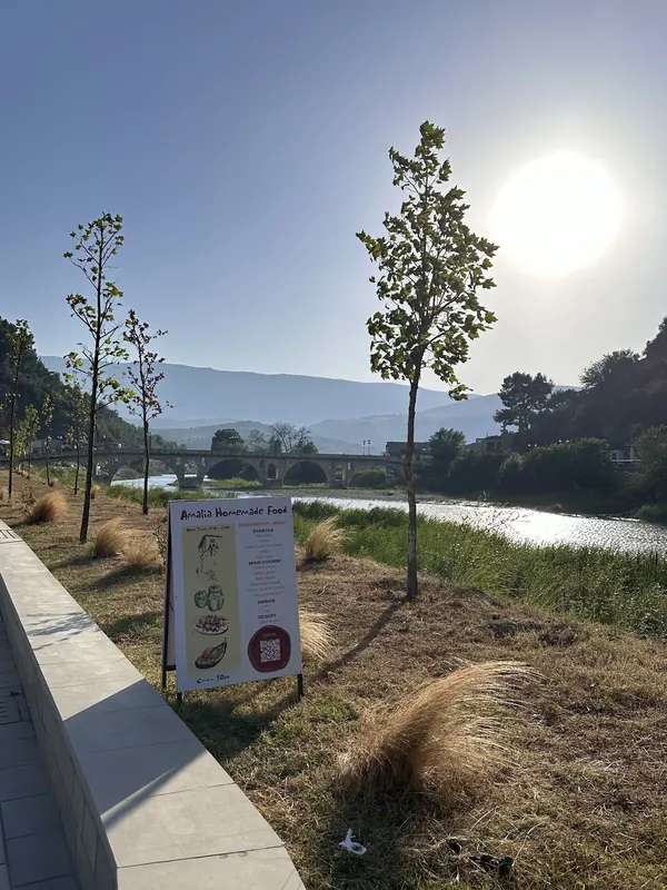 An Amalia Homemade Food menu board beside the Osum river, the old arched bridge and mountains behind