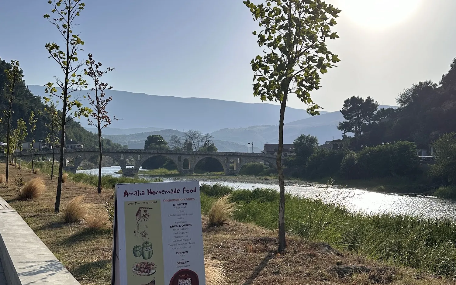 The Osum river in Berat at sunset, an old stone arched bridge and mountains behind, with an Amalia Homemade Food menu board on the riverside